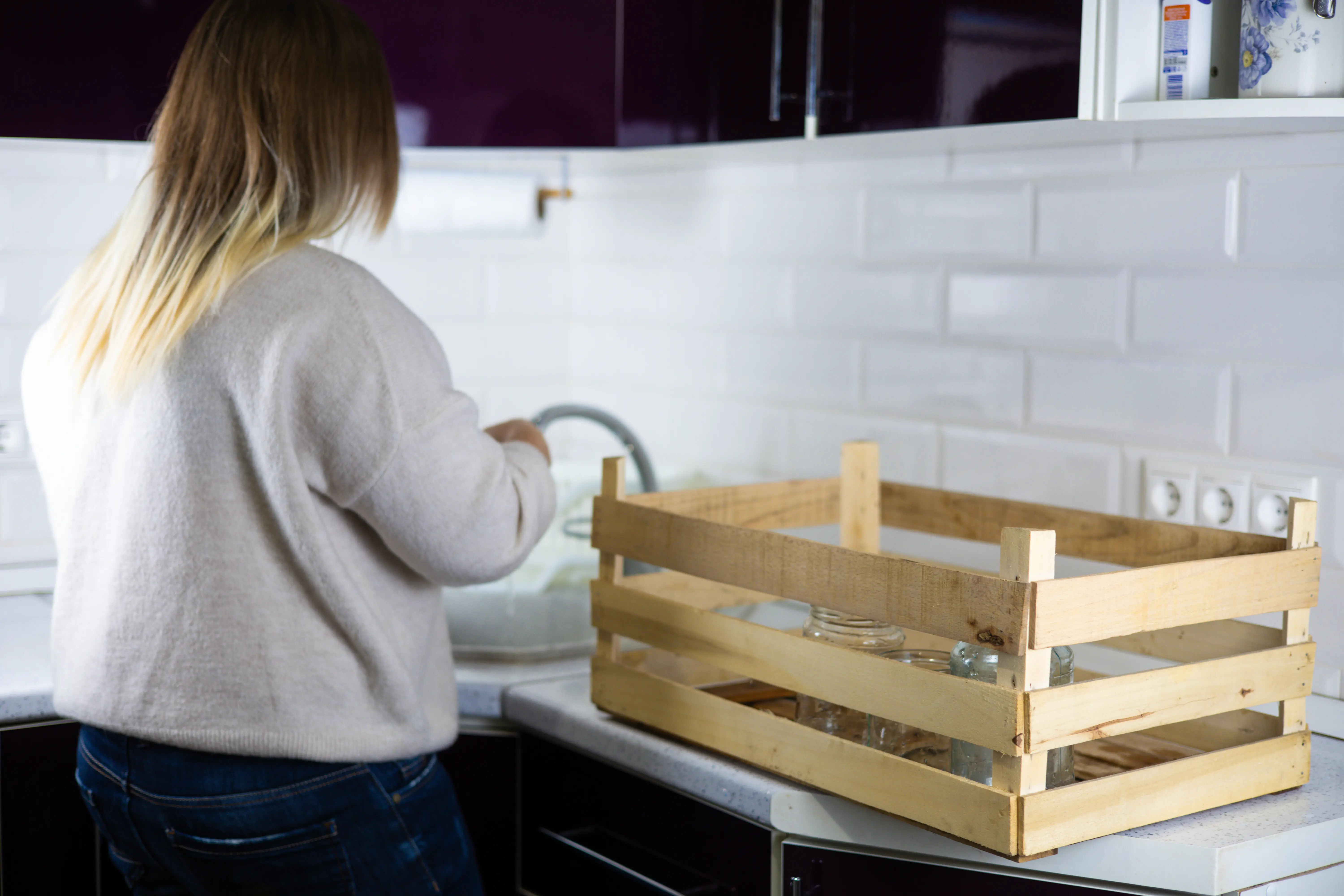 Eine Frau wäscht Glasbehälter und legt sie zum Recycling in eine Holzkiste. In der Küche sortiert sie Glasabfälle, um sie zu recyceln. Konzept sozialer Verantwortung und Umweltfreundlichkeit.